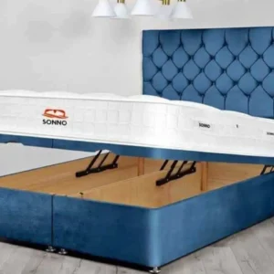 A contemporary beige upholstered Ottoman storage bed with the mattress base lifted by a hydraulic gas-lift mechanism, revealing a large internal storage space.