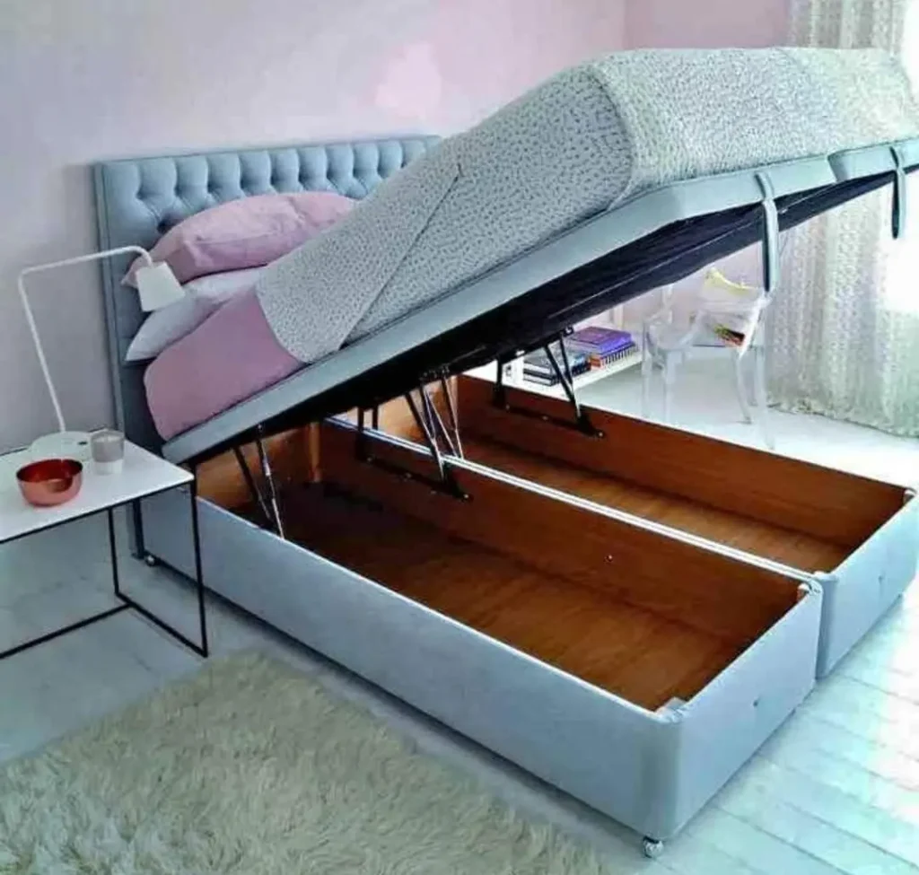 A contemporary beige upholstered Ottoman storage bed with the mattress base lifted by a hydraulic gas-lift mechanism, revealing a large internal storage space.