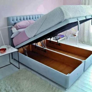 A contemporary beige upholstered Ottoman storage bed with the mattress base lifted by a hydraulic gas-lift mechanism, revealing a large internal storage space.